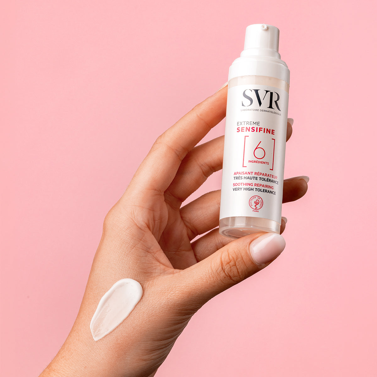 Hand holding a white SVR Sensifine Extrême pump bottle with a dab of white cream on the palm against a pink background.