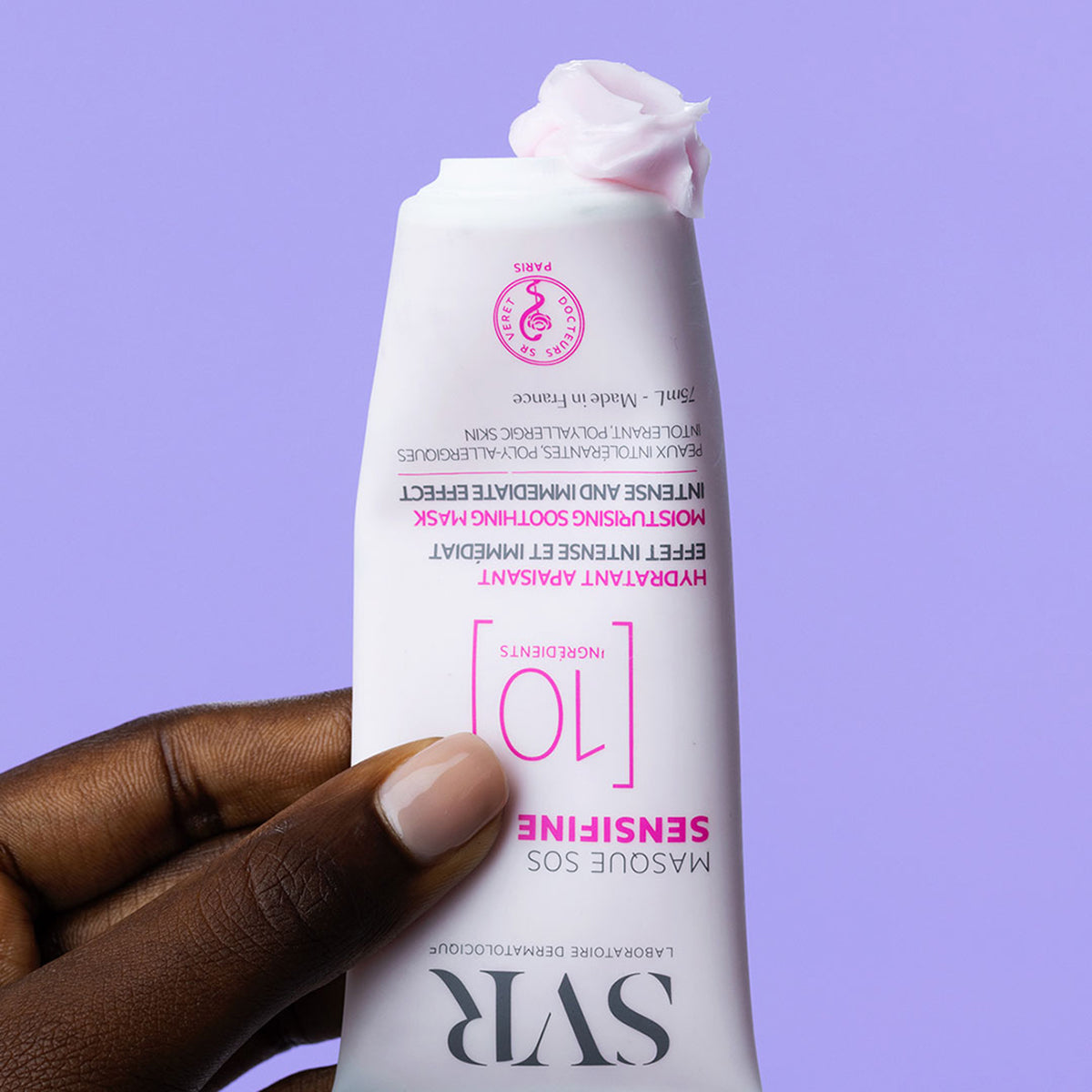 Hand holding a white 75ml SVR Sensifine SOS mask tube with a dollop of pale pink cream against a purple background.