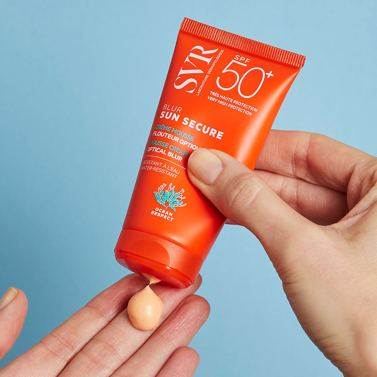 SVR SUN SECURE Blur SPF50+ orange tube dispensing a peach-colored mousse cream onto a hand against a blue background.