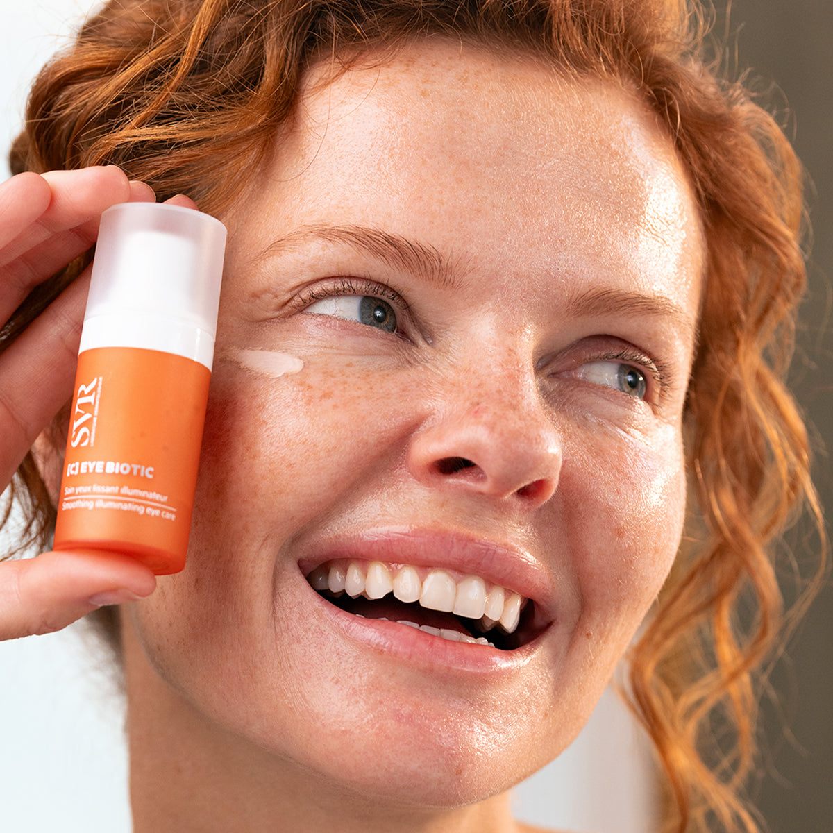 Woman holding an orange SVR C-EYE BIOTIC pump bottle while applying smoothing eye cream to her under-eye area.