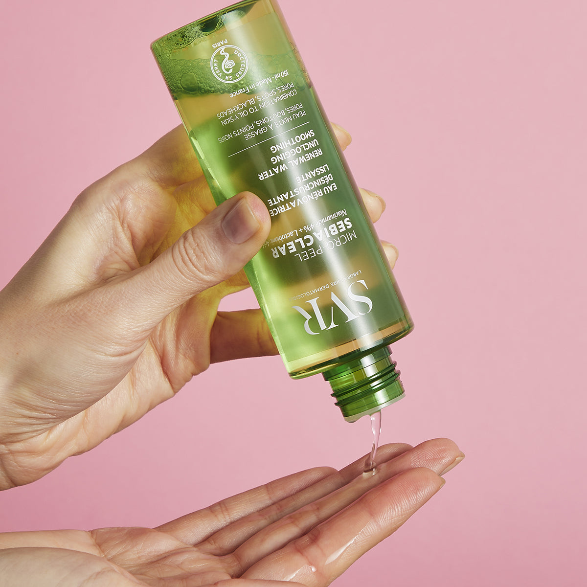 Flacon transparent verde SEBIACLEAR Micro-Peel turnând esență lichidă în palmă pe fundal roz.