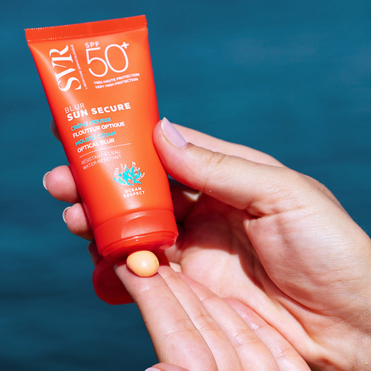 SVR SUN SECURE Blur SPF50+ orange squeeze tube with a dollop of mousse cream being applied to a hand against a blue background.