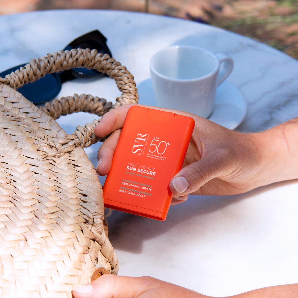 Spray solar portabil SVR Sun Secure SPF50+ cu ambalaj portocaliu compact, ținut în mână lângă o geantă de paie pe o masă albă.