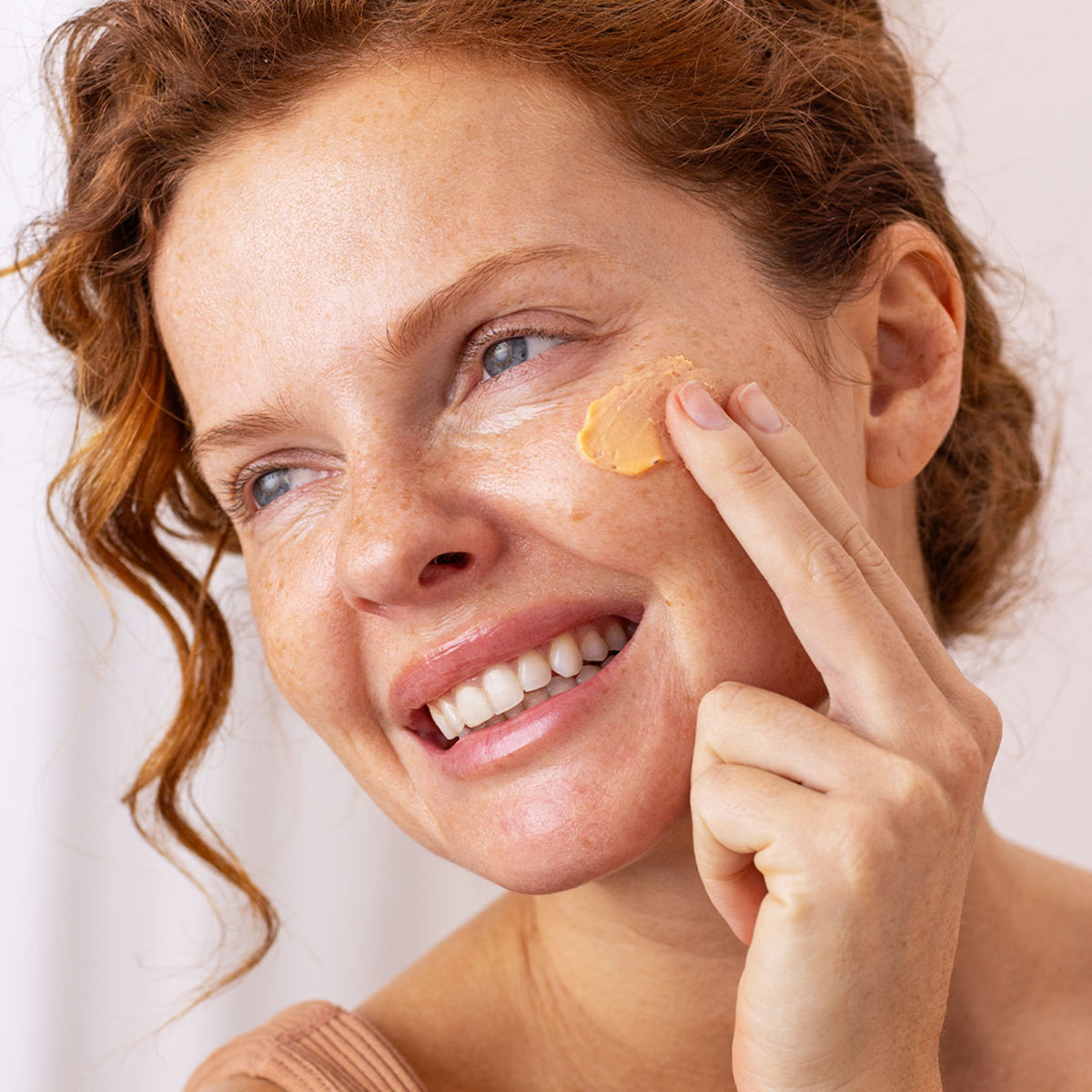 Smiling woman with red hair applying a peach-toned blurring SPF cream to her cheek against a neutral background.