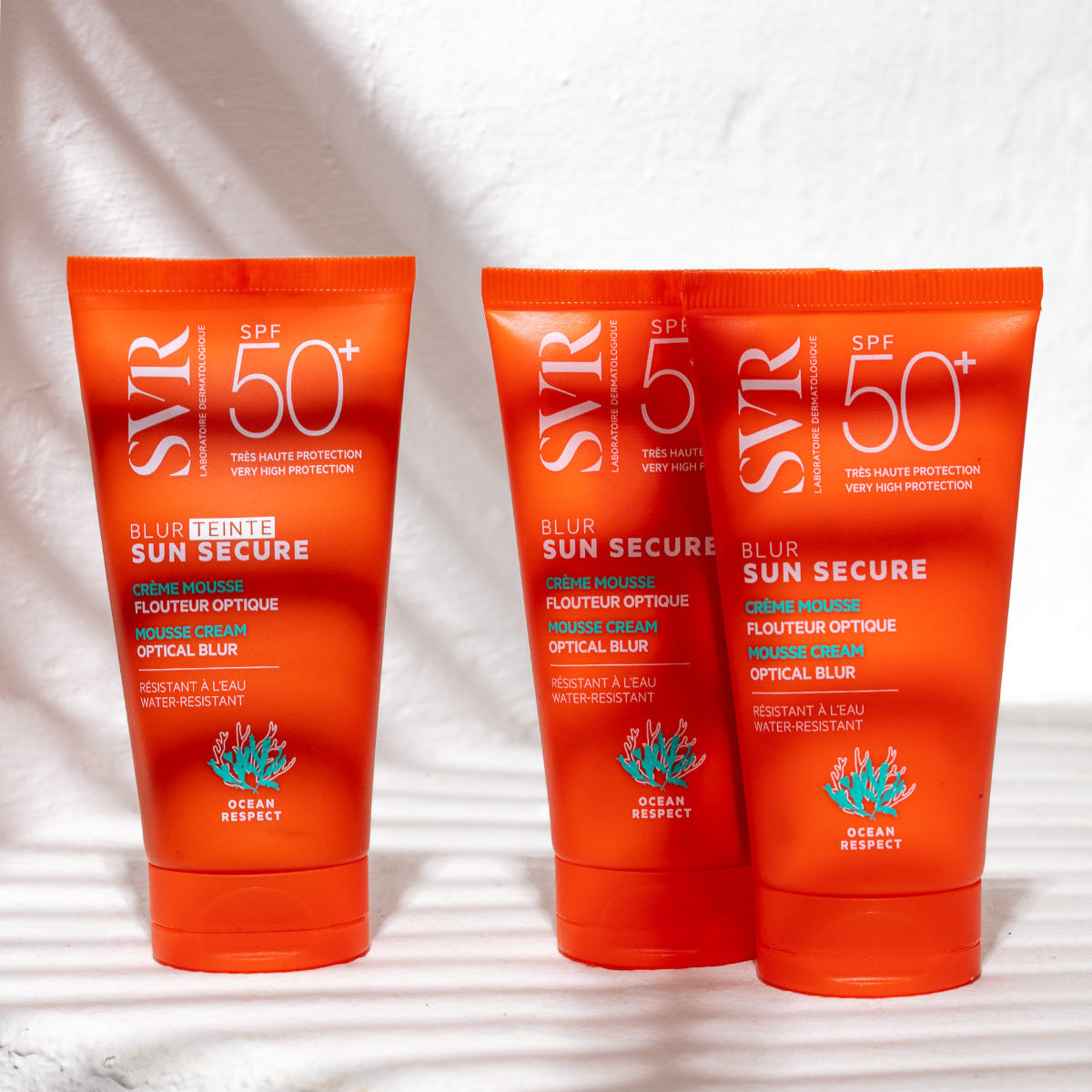 Three orange SVR SUN SECURE Blur SPF50+ mousse cream tubes standing on a white surface with soft palm leaf shadows.