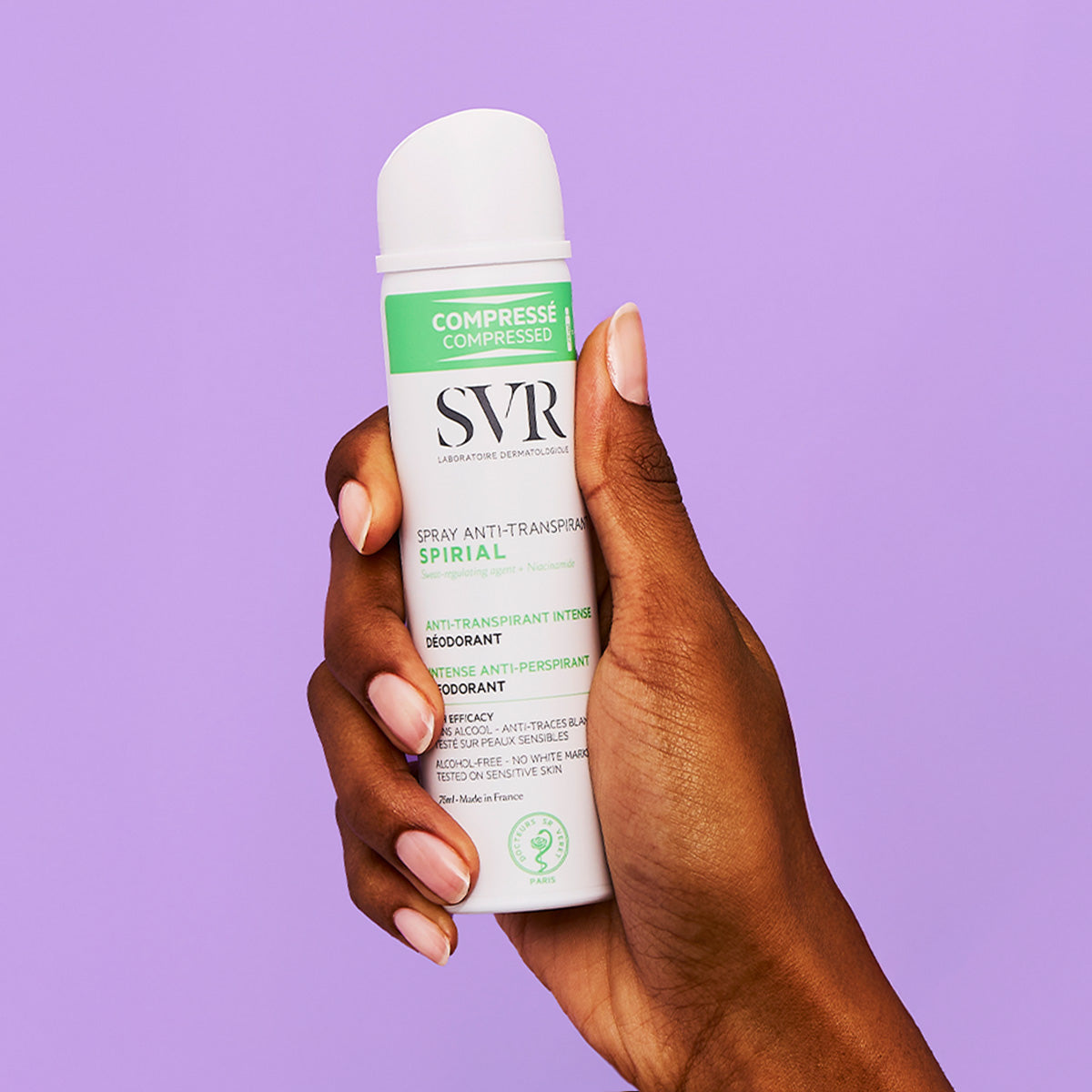 Mână ținând un spray antiperspirant SVR Spirial alb cu accente verzi pe fundal mov, ambalaj comprimat pentru călătorii.