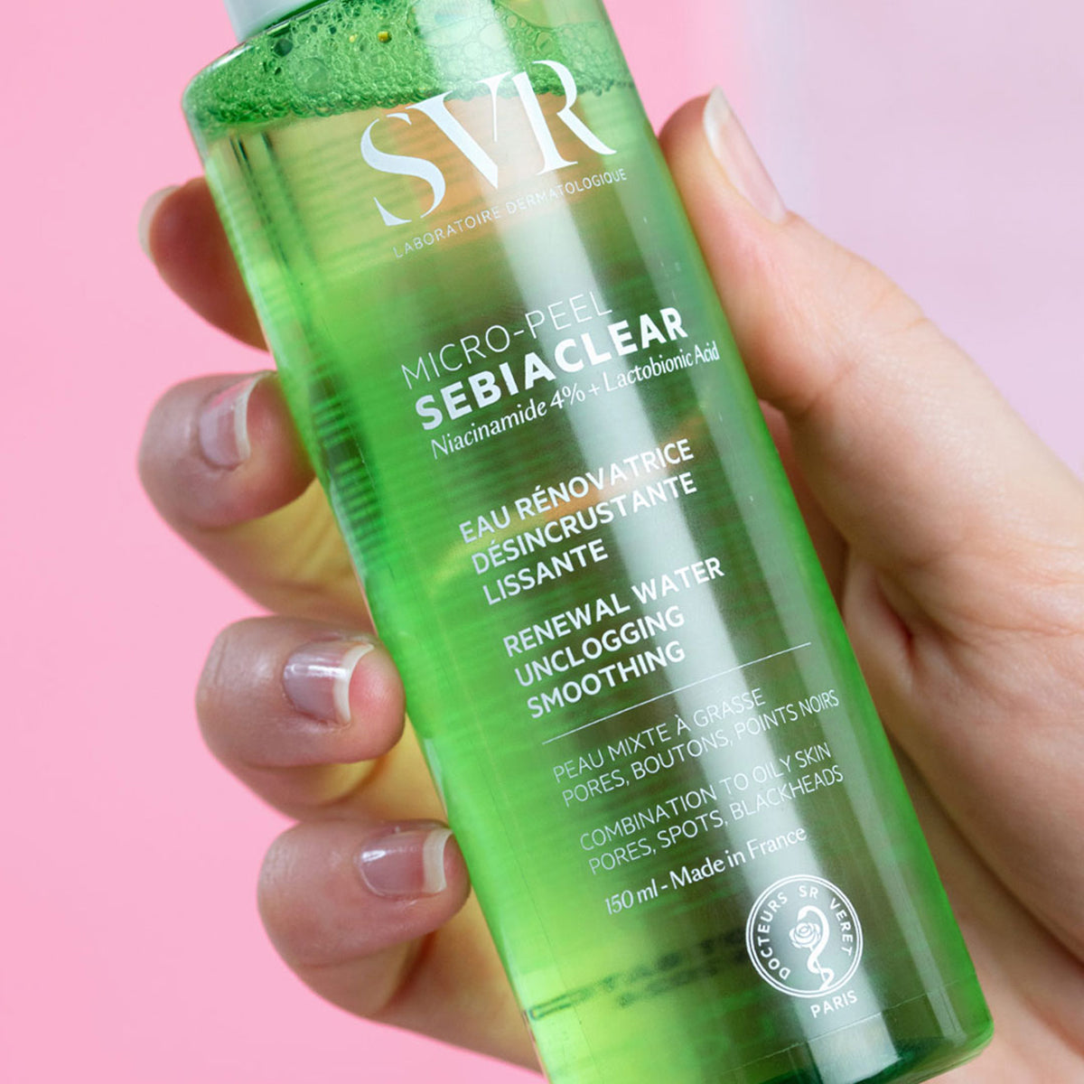 Flacon transparent de 150 ml cu loțiune verde SVR Sebiaclear Micro-Peel ținut în mână pe fundal roz.