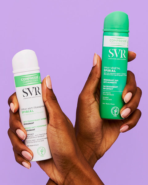 Two hands holding white and green SVR Spirial deodorant spray cans against a purple background.