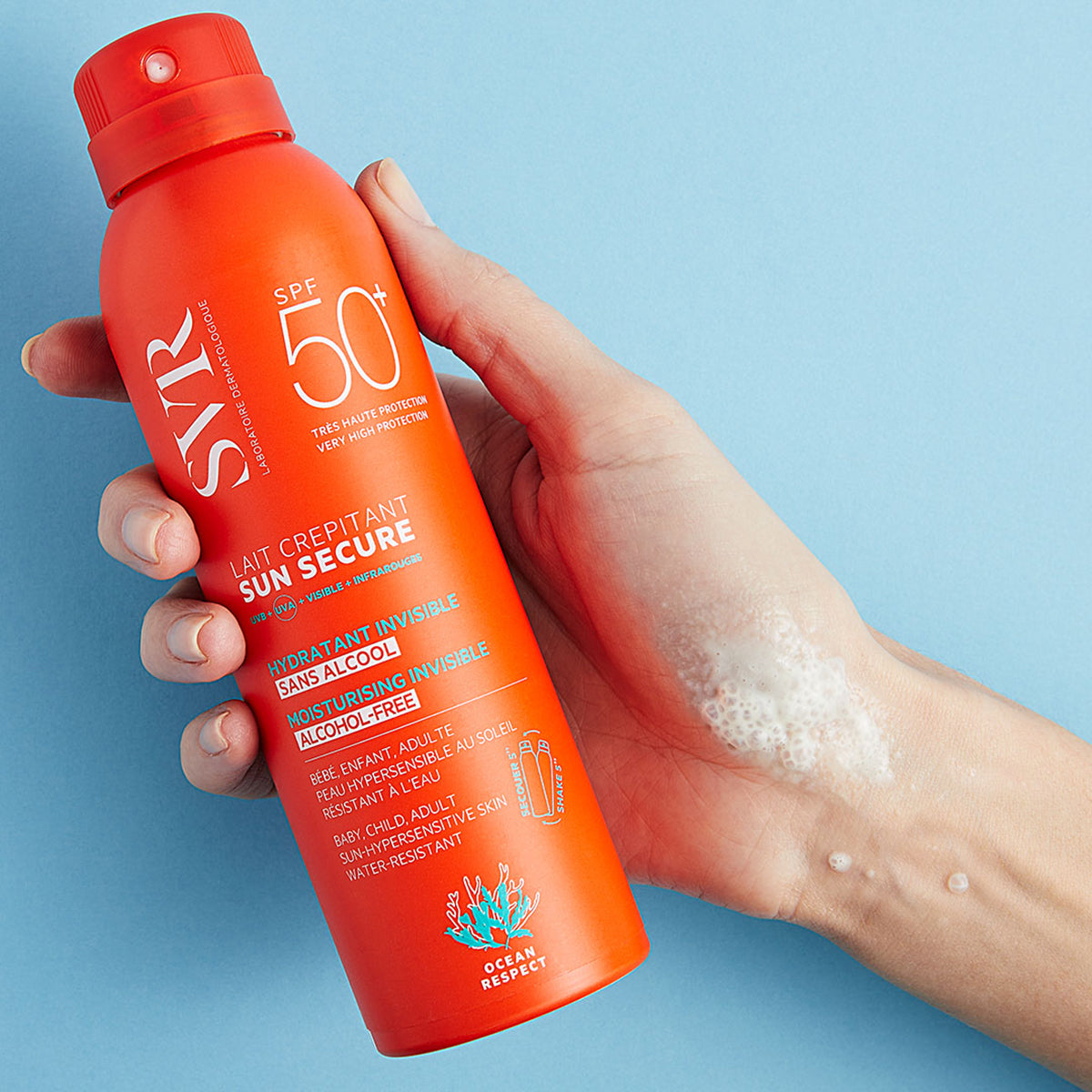 Spray solar SVR Sun Secure SPF50+ portocaliu, aplicat pe mână sub formă de loțiune spumoasă, pe fundal albastru deschis.