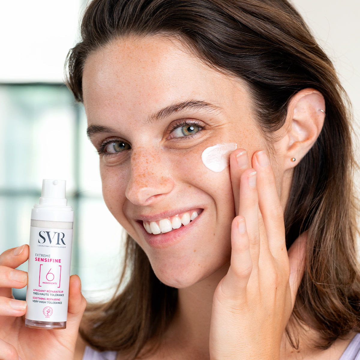 Smiling woman applying white SVR Sensifine Extrême cream from a pump bottle to her cheek.