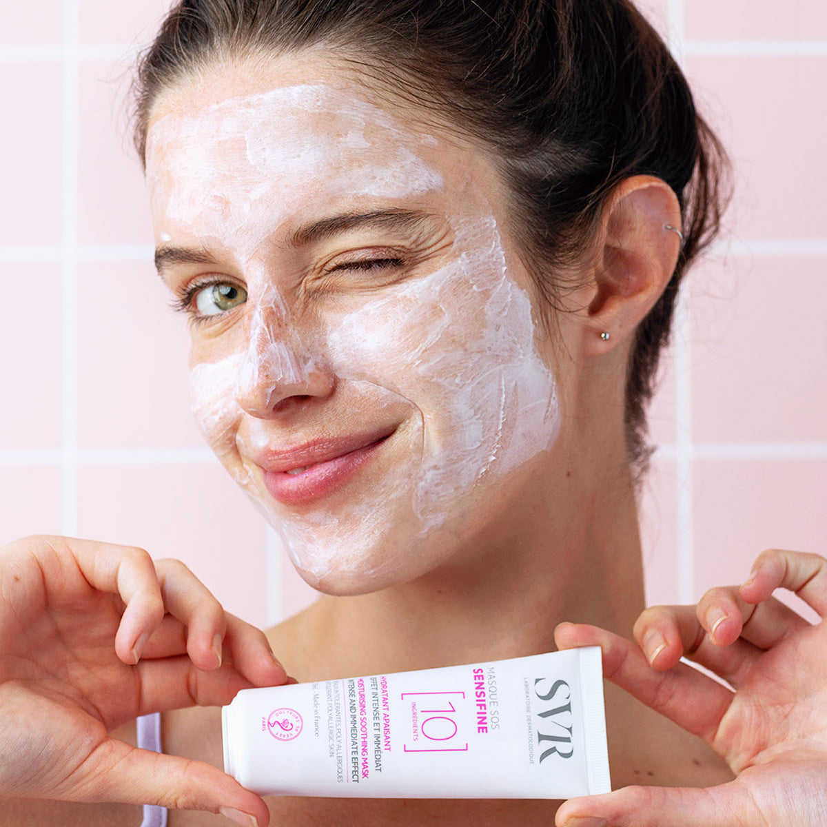 Woman winking with white SVR Sensifine SOS soothing face mask applied, holding the white product tube against a pink tile wall.