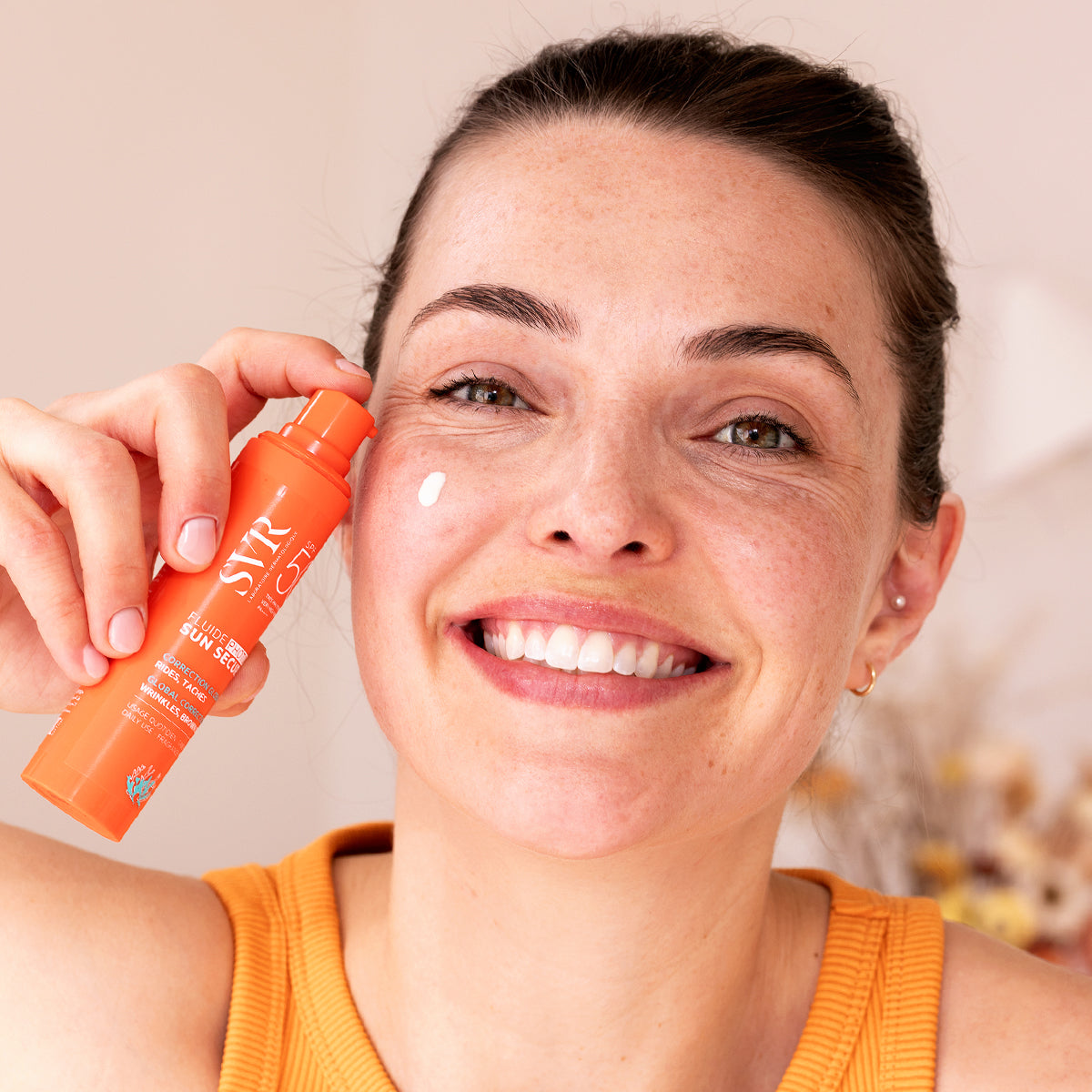Smiling woman holding an orange SVR SUN SECURE SPF50+ pump bottle with a dab of sunscreen on her cheek.