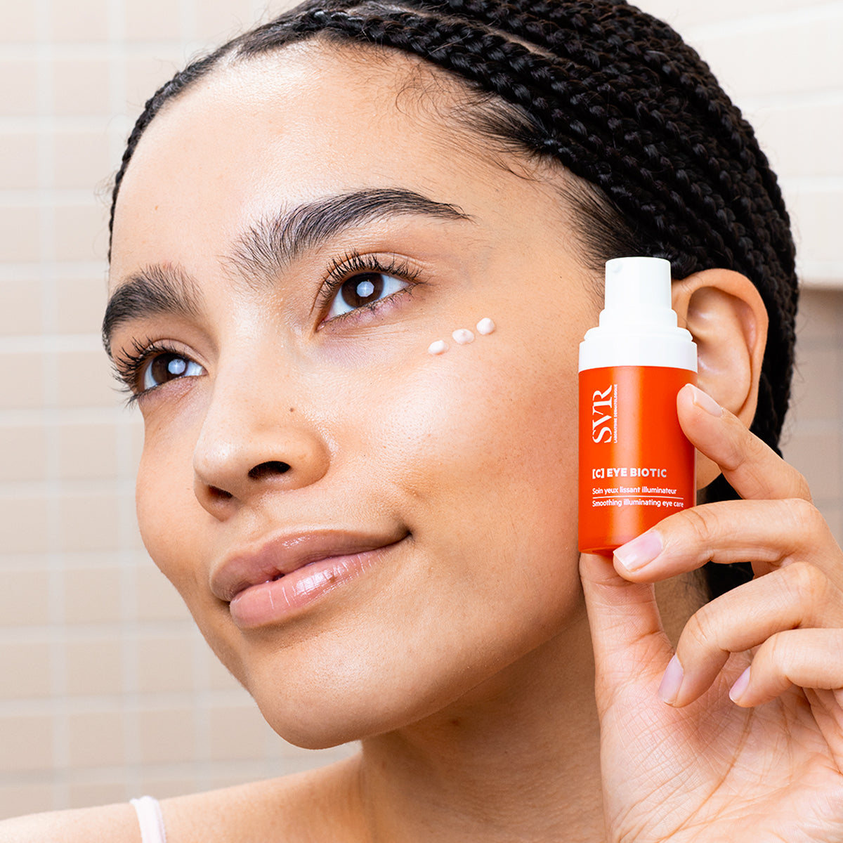Woman holding an orange SVR C-EYE BIOTIC pump bottle with cream dots applied under her eye.