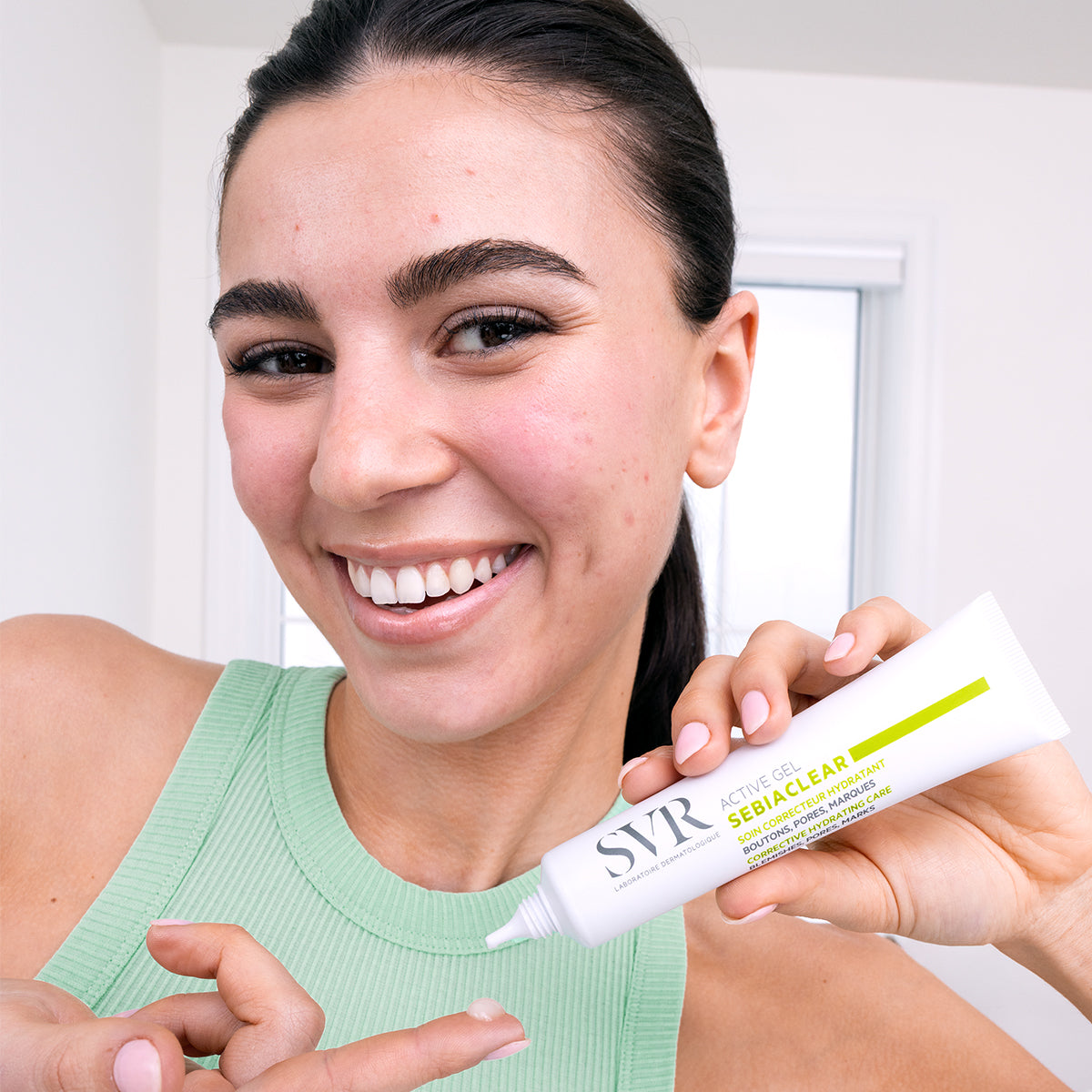 Smiling woman holding a white tube of SVR Sebiaclear Active Gel and applying a small amount to her fingertip.