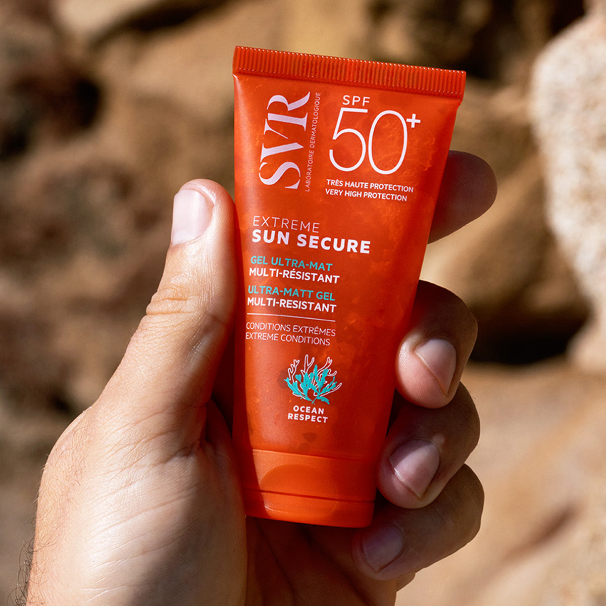 Tub portocaliu de cremă solară SVR Sun Secure SPF50+ ținut în mână, cu text alb și logo Ocean Respect pe fundal stâncos.