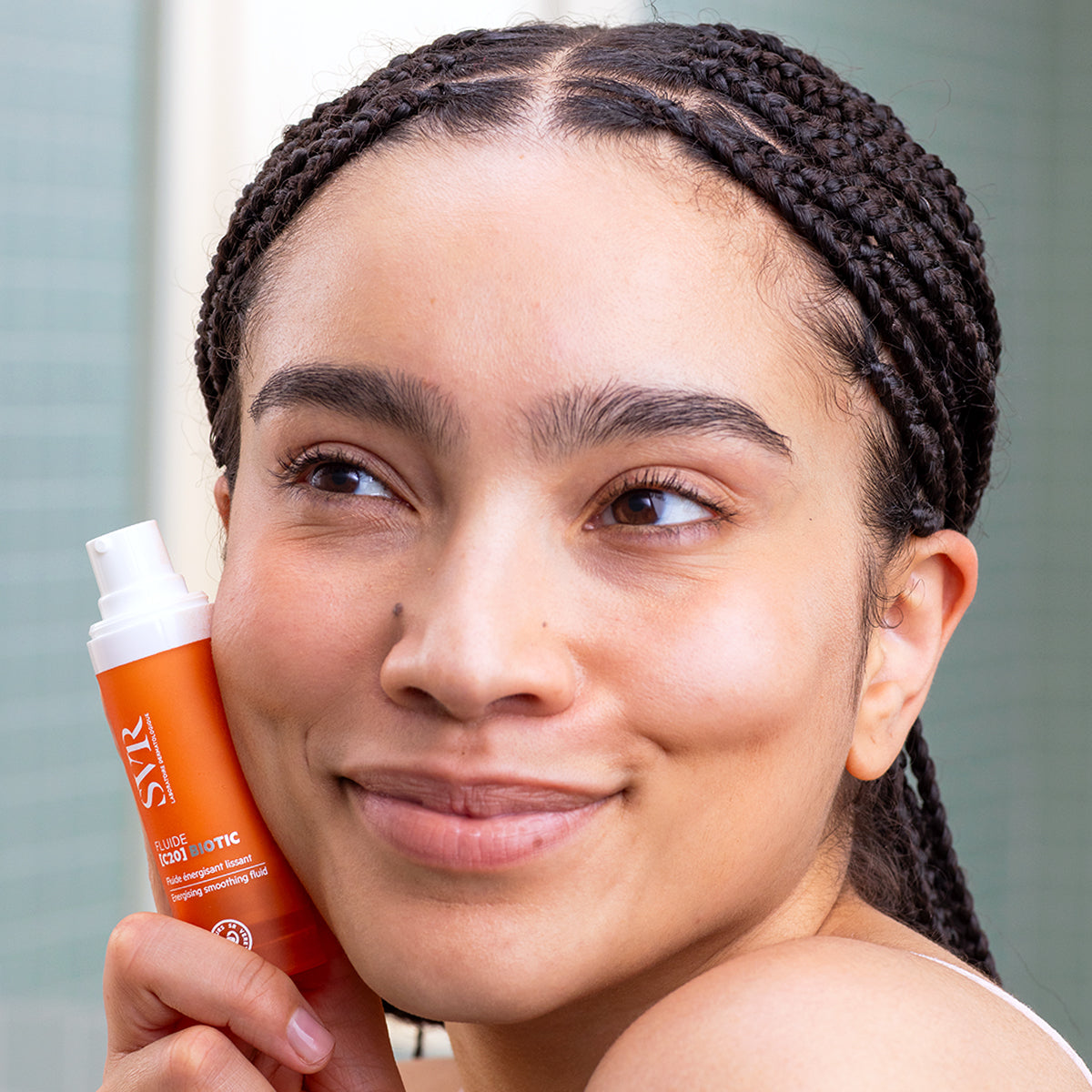 Woman holding an orange SVR Fluide [C20] BIOTIC pump bottle against her cheek with a radiant, glowing complexion.