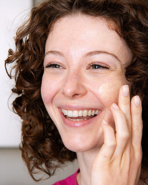 Smiling woman with curly hair applying tinted soothing cream to her cheek.
