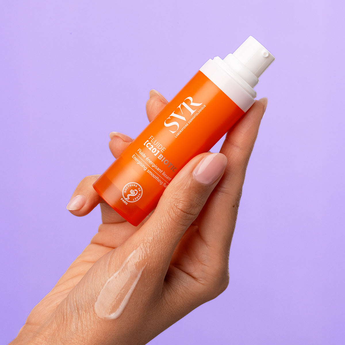 Hand holding an orange SVR Fluide [C20] BIOTIC pump bottle with a drop of clear smoothing fluid on the skin.
