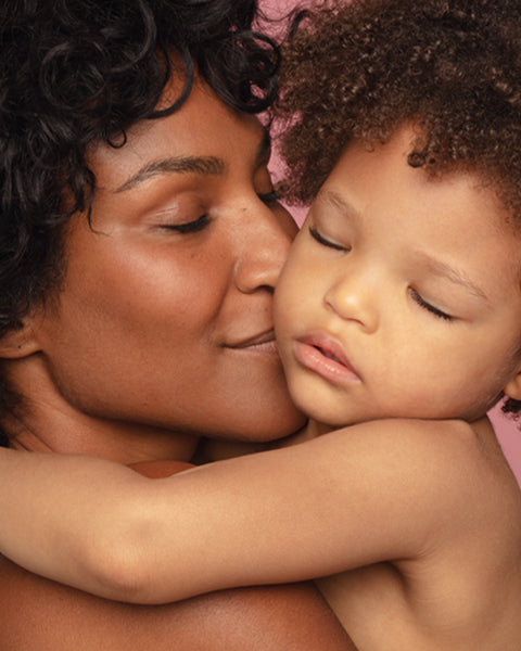 A Black mother with curly hair gently embracing and kissing her young child against a soft pink background.